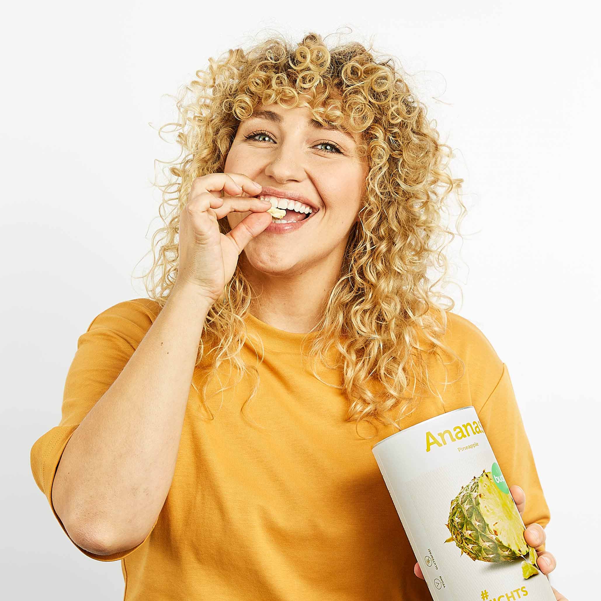 Junge Frau mit blonden Locken hat eine Dose buah Ananas in der Hand und isst nebenher gefriergetrocknete Ananas