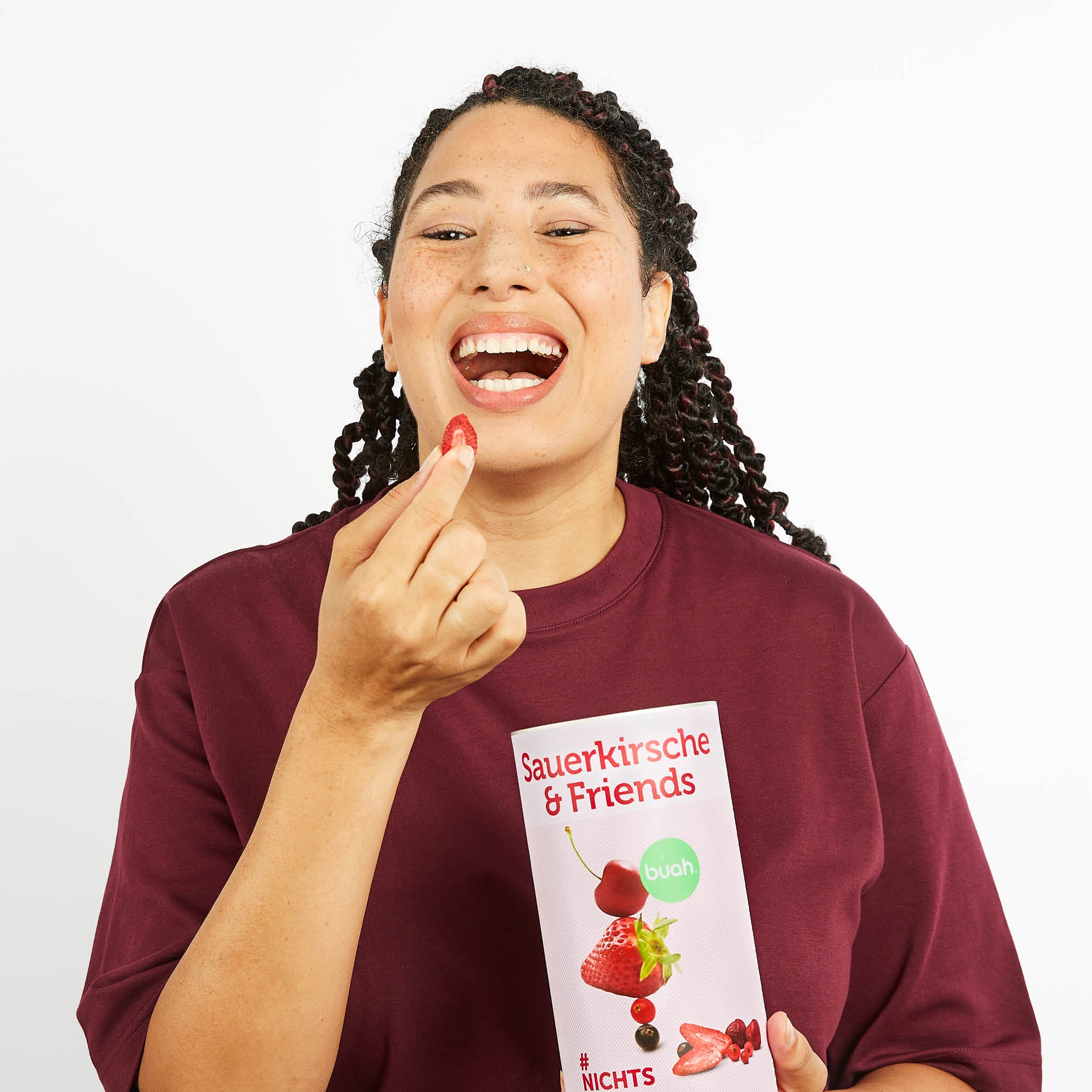 Lachende Frau mit Locken und dunkelrotem Shirt hat eine Dose buah Sauerkrische & Friends in der Hand und isst die Fruechte