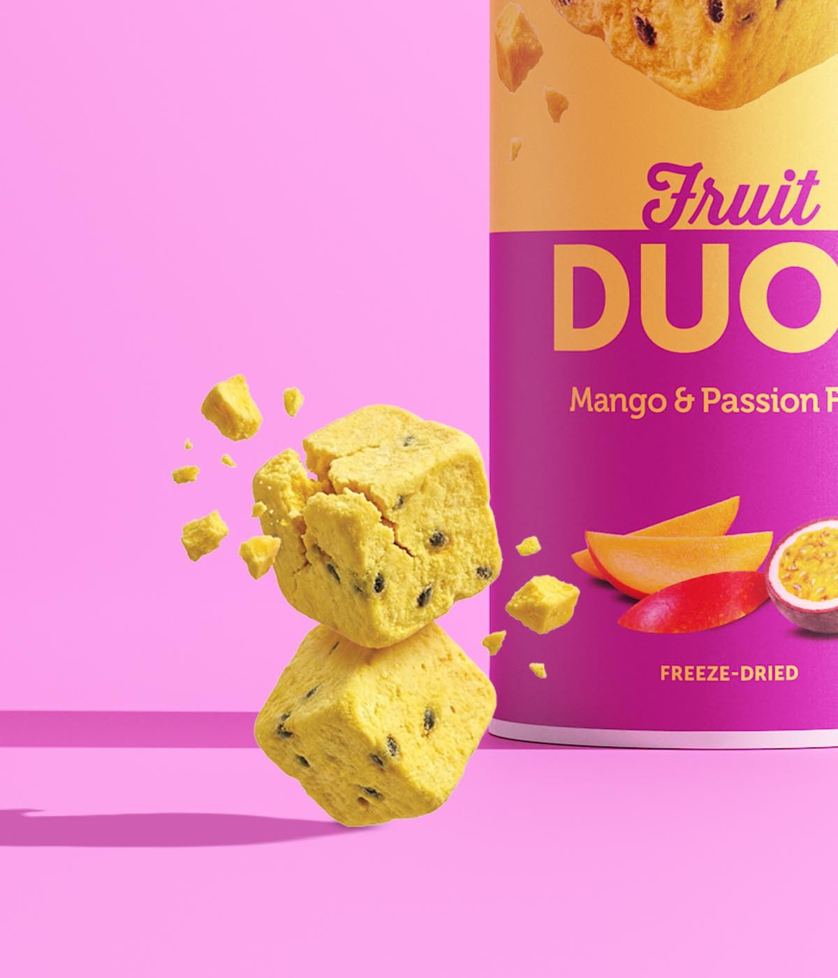 Ein buah gefriergetrockneter Fruchtwürfel der Sorte Mango & Maracuja zerbricht in der Luft. Darunter schwebt ein zweiter intakter Fruchtwürfel. Der Hintergrund ist rosafarben.