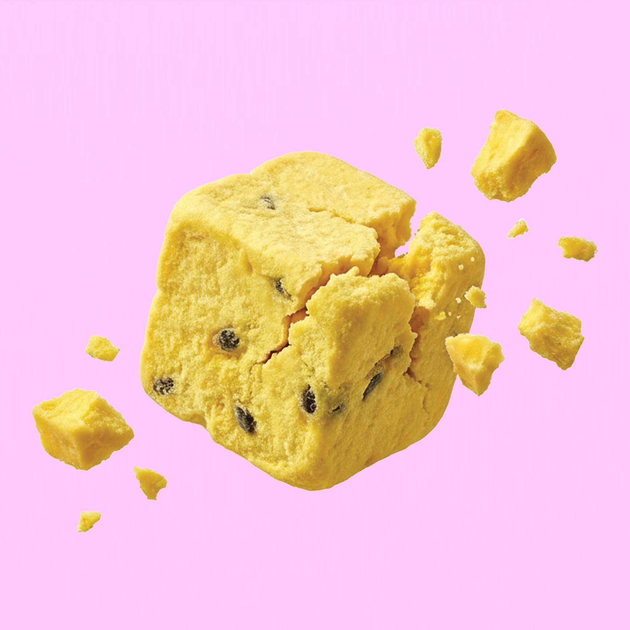 Ein quadratischer, buah gefriergetrockneter Fruchtwürfel der Sorte Mango & Maracuja zerbricht in der Luft vor einem rosafarbenen Hintergrund. Kleine gelbe Stückchen und dunkle Maracujakerne fliegen davon.