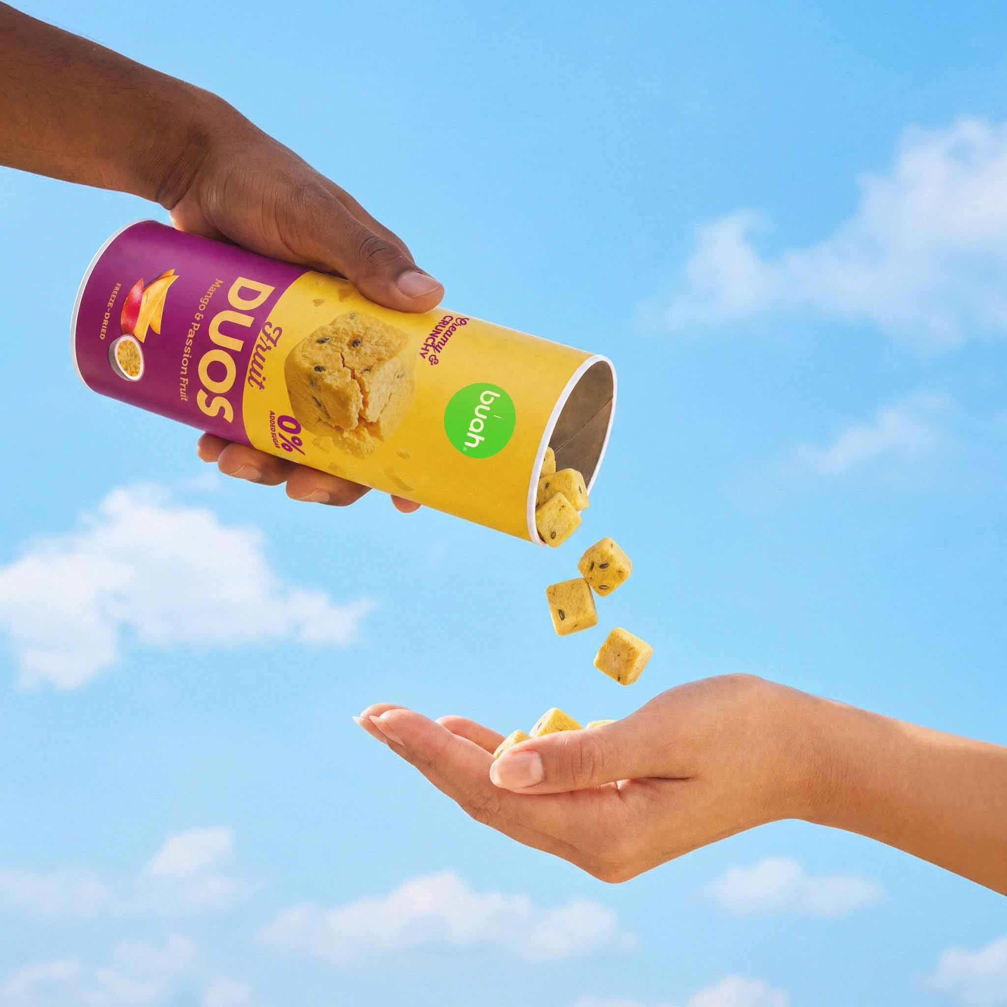 Eine Hand schüttet quadratische, buah gefriergetrocknete Fruchtwürfel der Sorte Mango & Maracuja aus einer Slim-Dose in die geöffnete Handfläche einer zweiten Person. Im Hintergrund sieht man blauen Himmel und ein Picknicktuch.