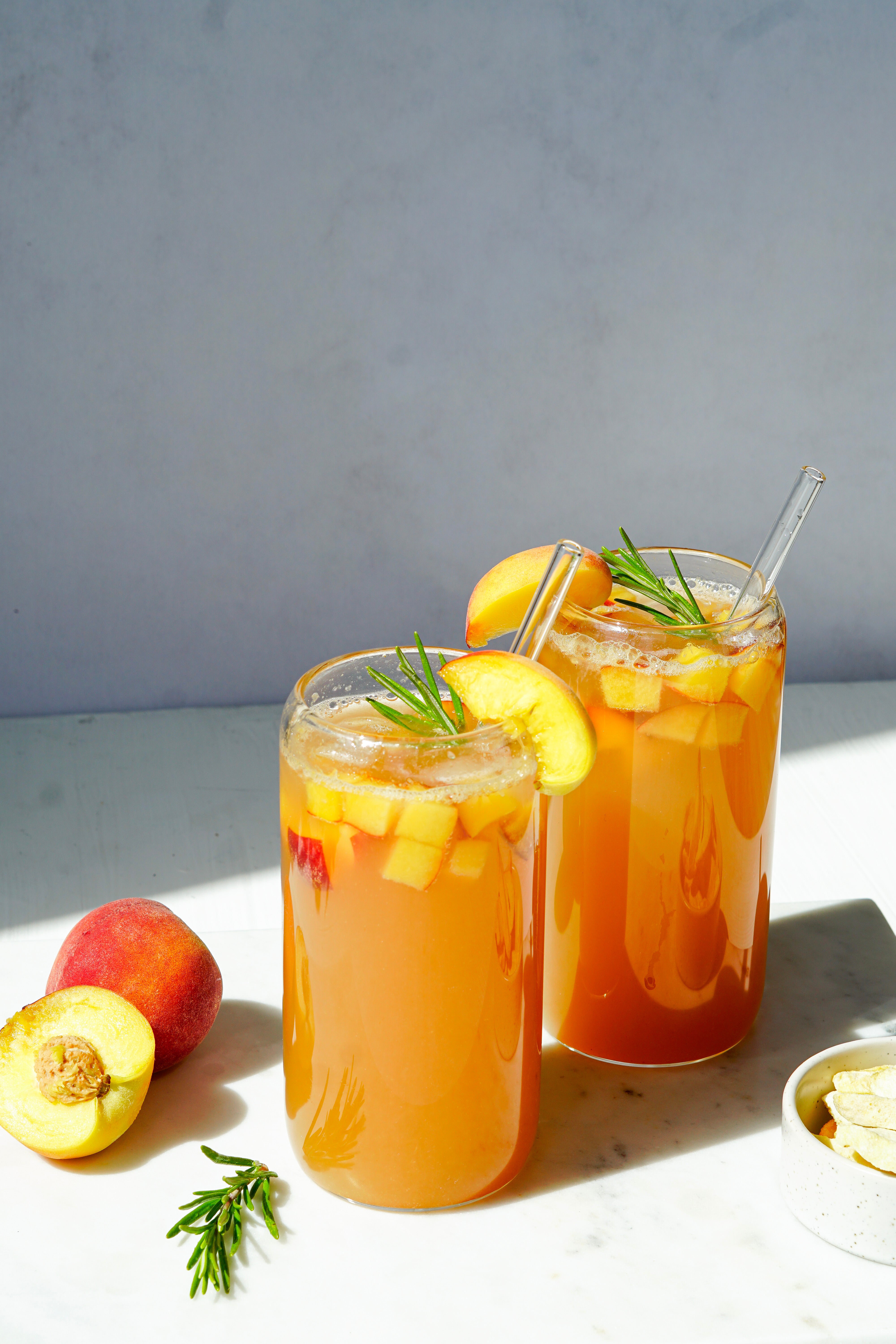 Ein erfrischendes Sommergetränk. Zwei Gläser gefüllt mit Pfirsich-Eistee, frischen Pfirsichstücken, Eiswürfeln, Rosmarin und Glasstrohhalmen stehen im Sonnenlicht. Daneben liegen eine aufgeschnittene frische Pfirsichhälfte und ein Schälchen mit buah gefriergetrockneten Früchten.