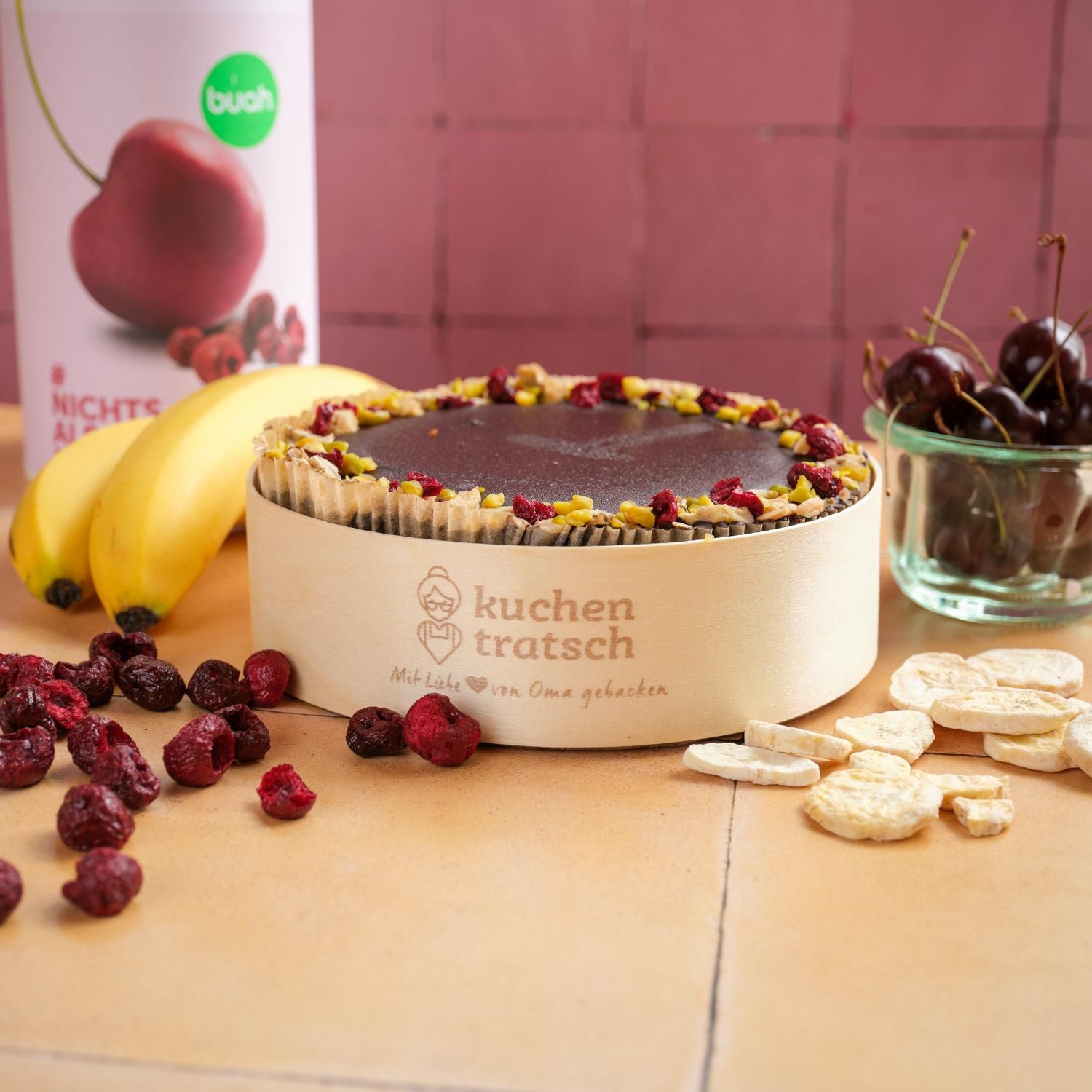Kuchen mit buah gefriergetrockneten Fruechten gebacken von Kuchentratsch in einer Holzform mit Kuchentratsch Logo, buah Sauerkirsche und Banane im Hintergrund