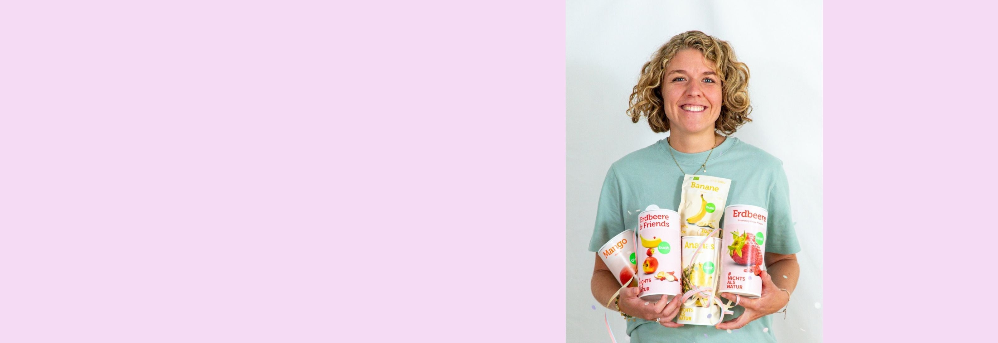 Jessica Gruenderin von buah in gruenem buah Shirt mit buah gefriergetrockneten Fruechten in der Dose im Arm - Konfetti und Luftschlangen vor rosa Hintergrund  