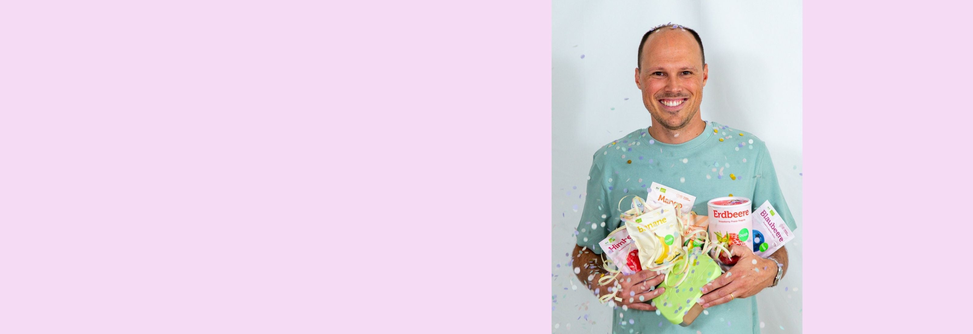 Tobi von buah im gruenem buah Shirt mit buah gefriergetrockneten Fruechten in der Dose im Arm - Konfetti und Luftschlangen vor rosa Hintergrund