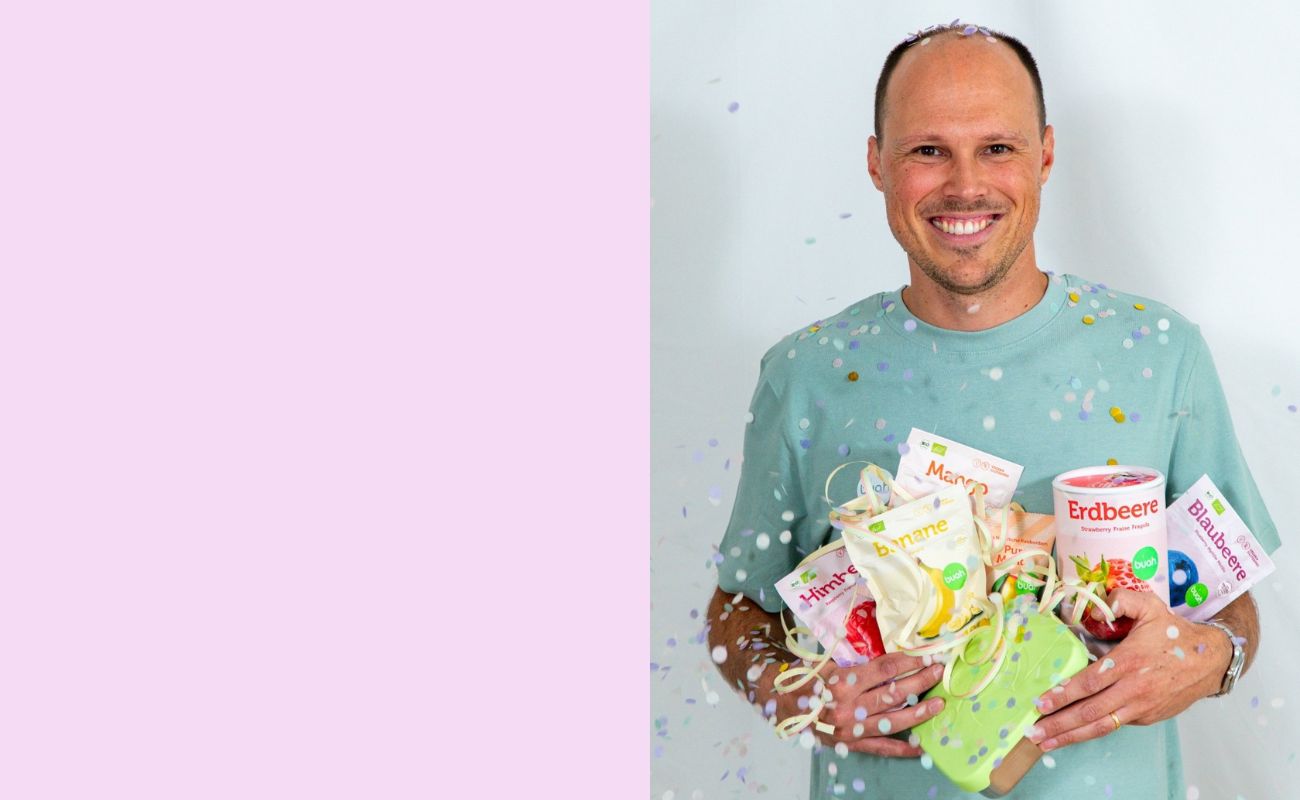 Tobi von buah im gruenem buah Shirt mit buah gefriergetrockneten Fruechten in der Dose im Arm - Konfetti und Luftschlangen vor rosa Hintergrund