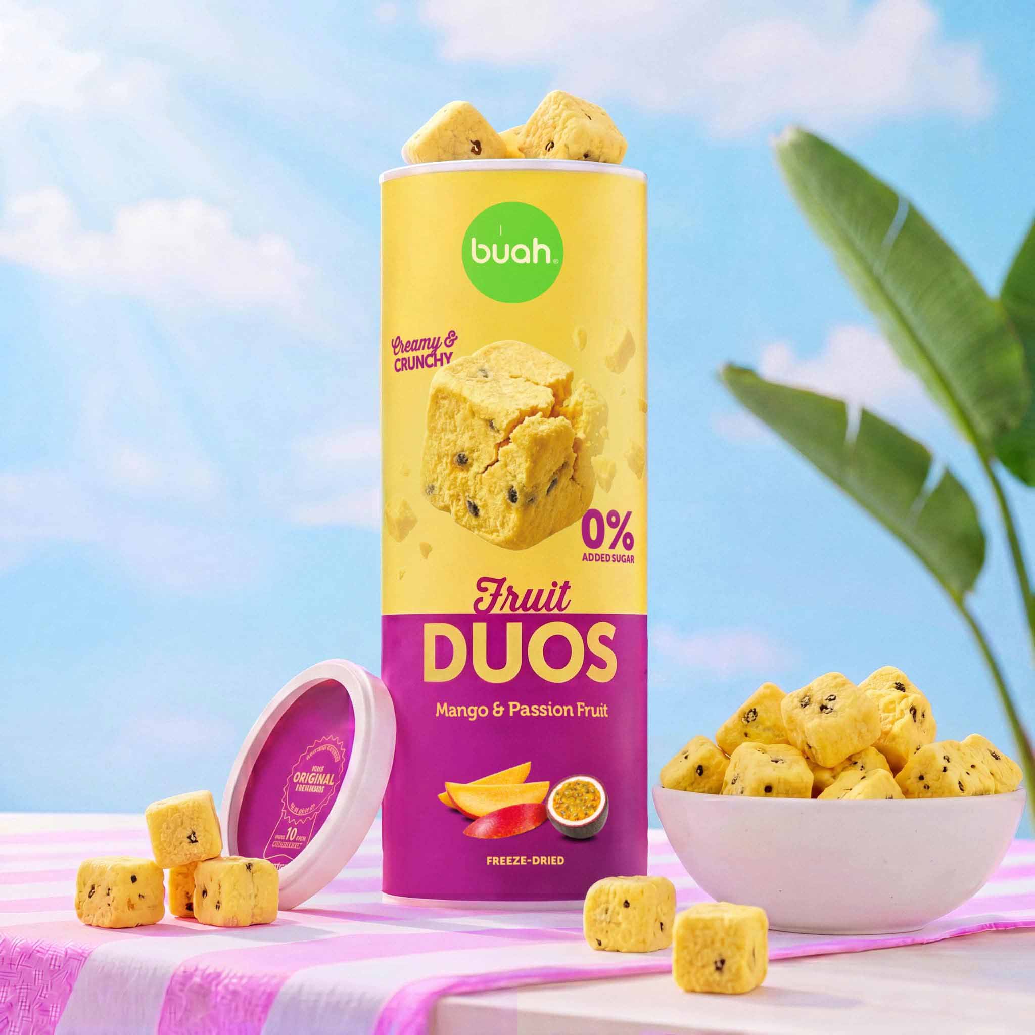 Eine hohe Slim-Dose buah Fruit DUOS Mango & Passion Fruit steht geöffnet vor einem strahlend blauen Himmel mit ein paar Wolken. Im Hintergrund sieht man die großen grünen Blätter einer Bananenpflanze. Rechts steht eine Schale mit Fruchtwürfeln.